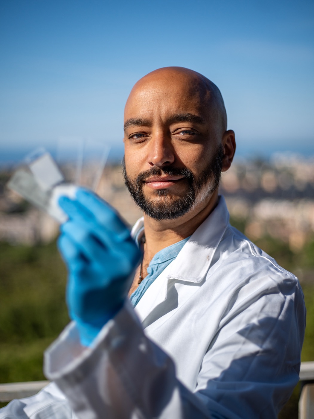 Fred Dante David outdoors in a lab coat and blue gloves, with the Genova skyline in the background