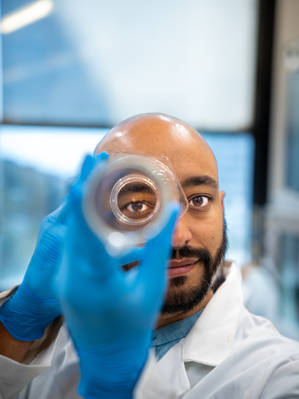 Fred Dante David in a lab coat and blue gloves, holding a small sample vessel up to his face in the laboratory